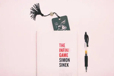 A book titled 'The Infinite Game' by Simon Sinek with a bookmark and a pen on a pink background. 