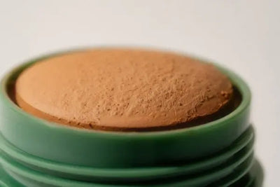 A close-up of a brown makeup stick with a textured surface in a green cylindrical container. The soft lighting and neutral background highlight the product's earthy tones and natural appeal.