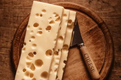 Slices of Swiss-style cheese with holes on a rustic wooden cutting board, accompanied by a cheese knife engraved with 