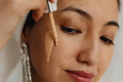 A close-up of a person applying liquid concealer with a dropper to their cheek. The medium-toned skin, silver earring, and soft lighting emphasize a beauty routine in progress.