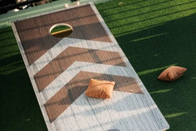 Cornhole board on green artificial turf with a faux wood grain and chevron pattern. One orange-brown beanbag sits on the board; another rests on the grass, suggesting an outdoor game in progress.
