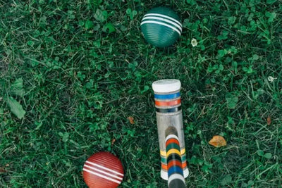 Top-down view of croquet equipment on grass. A mallet with colorful bands and two striped balls (green and red) rest on a natural lawn with clover and small flowers, evoking a casual outdoor vibe.