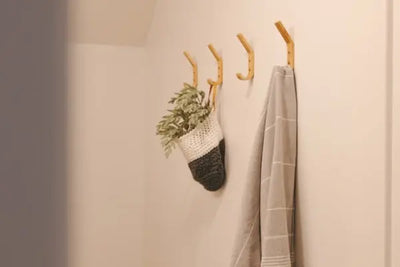 Light wooden hooks on an off-white wall, holding a folded striped towel and a two-toned hanging basket with green foliage. Minimalist, cozy decor with soft, warm lighting.