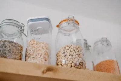 Close-up of pantry shelf with clear jars and a plastic container holding seeds, self-raising flour, chickpeas, and red lentils. Clip-top lids, light wood shelf, soft focus background.