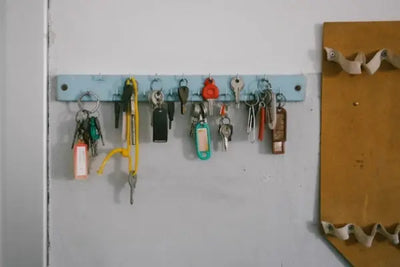 Light blue key rack on a gray wall holding various keys and colorful keychains, including red, yellow, and peach accents. Adjacent is a brown panel with white wavy elements, adding a decorative touch.