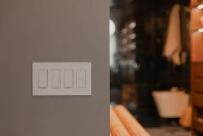 White four-switch light panel on a light grayish-brown wall, with a blurred interior background featuring a towel, white fixture, and wooden elements. Modern, clean design in a cozy home setting.