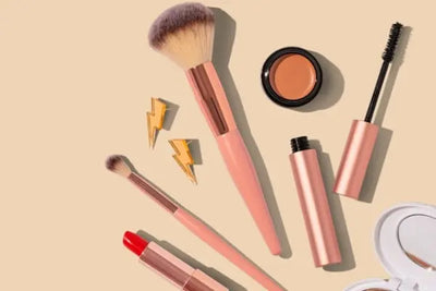 A flat lay of makeup essentials on a beige background, featuring rose gold brushes, red lipstick, mascara, a concealer pot, a compact mirror, and gold lightning bolt accessories for a chic aesthetic.