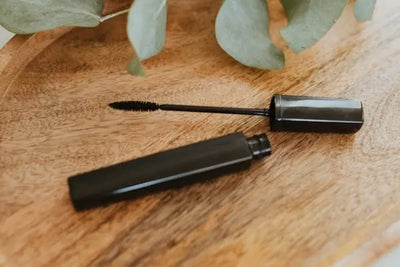 An open black mascara tube and wand rest on a textured wooden surface, with blurred green leaves in the background. The soft lighting highlights the sleek design and natural, minimalist aesthetic.