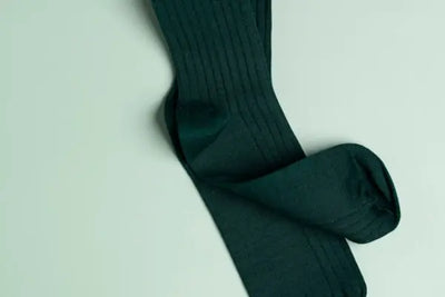 Dark green ribbed men’s socks laid flat on a light mint background. Soft, even lighting highlights the texture and overlapping fold, conveying a clean, minimalist look and cozy, classic style.