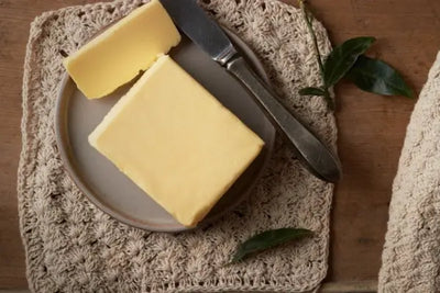 Top-down view of a small plate with a block and wedge of pale yellow butter, a vintage butter knife, on a beige crocheted mat over a wooden table, with a couple of small green leaves nearby.