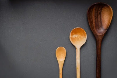 Top-down view of three wooden serving spoons in descending size, arranged vertically on a dark textured background. The spoons vary in wood tone from pale to medium to dark, highlighting natural grain.