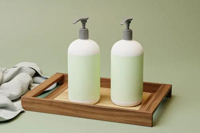 Two light green and white pump bottles on a wooden tray with a soft gray towel, set against a solid light green background. Minimalist, spa-like scene suggesting soap, lotion, or bathroom accessories.
