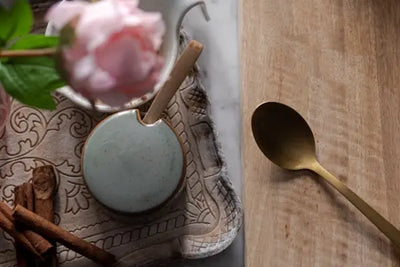 Overhead still life with a soft pink rose, bundled cinnamon sticks on a carved tray, a small blue-green dish holding a wooden scoop, and a gold spoon resting diagonally on a light wooden board.