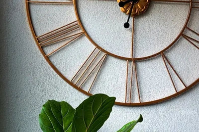 Golden Roman numeral wall clock on a textured light wall, with dark green Fiddle Leaf Fig leaves in the foreground. Elegant and natural elements combine for a stylish, timeless indoor decor vibe.