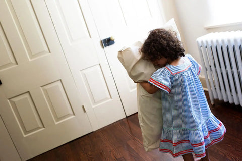 A young child in a blue striped dress hugs an American Blossom Linens Natural Cotton Bed Pillowcase, made from USA-grown cotton, in a bright room with wooden floors, cream double doors, and a white radiator.