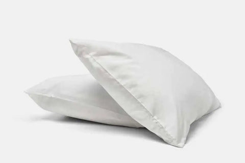 Two soft, white pillows are stacked on a plain white background, each covered in American Blossom Linens’ Natural Cotton Bed Pillowcases made from luxurious, USA-grown cotton. The covers look smooth and the pillows appear plush and fluffy.