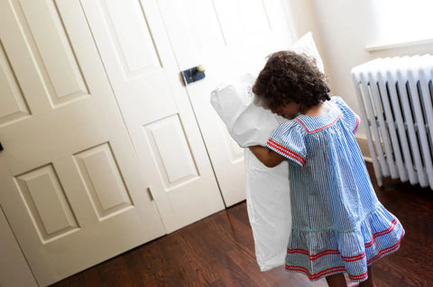 A young child in a blue striped dress stands by cream closet doors, holding American Blossom Linens’ Natural Cotton Bed Pillowcases made from luxurious USA-grown cotton, facing away from the camera near a radiator.
