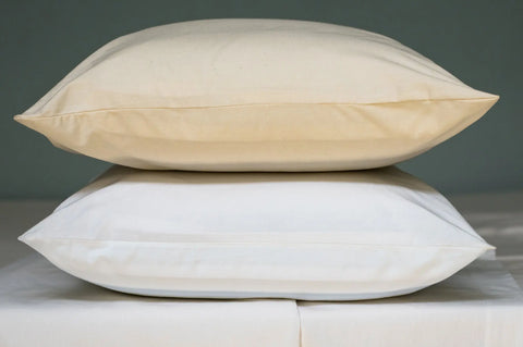 Two pillows are stacked, one in beige and one in white, each with luxurious Natural Cotton Bed Pillowcases by American Blossom Linens, made from USA-grown cotton, resting on a white surface against a plain background.
