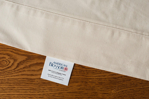 A close-up of Natural Cotton Bed Pillowcases by American Blossom Linens, made from 100% USA-grown organic cotton, shown with a white tag on wood that reads: Sustainably Made in the U.S.A. by your friends at Thomaston Mills.
