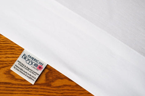 A close-up of Natural Cotton Bed Pillowcases by American Blossom Linens, featuring a tag that reads “American Blossom Linens, 100% USA grown organic cotton, Made in USA,” displayed on a wooden surface.