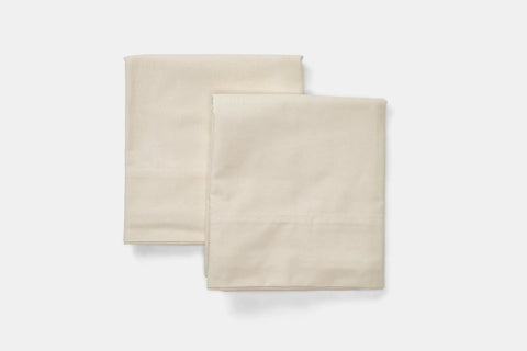 Two neatly folded Natural Cotton Bed Pillowcases by American Blossom Linens in light beige are placed side by side on a white background, adding a luxurious touch to your bedding.