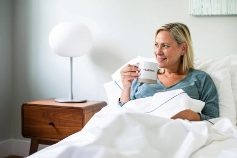 A woman with blonde hair sits up in bed, smiling and holding a THANKFUL mug. She is covered with American Blossom Linens’ Cotton Piping Design Bed Sheet Set, and a wooden nightstand with a lamp stands beside her.