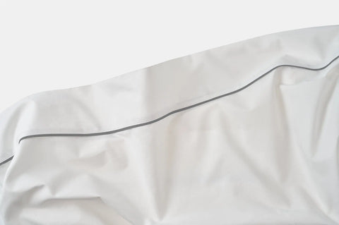 A close-up of the Cotton Piping Design Bed Sheet Set by American Blossom Linens, featuring USA-grown cotton with a slim dark gray trim running diagonally across the white fabric, highlighting gentle wrinkles and folds.