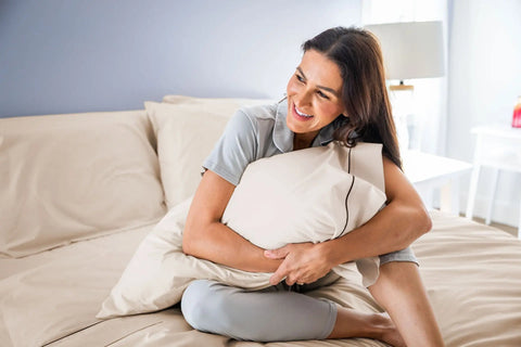 A smiling woman in light gray pajamas hugs a pillow on a neatly made bed with American Blossom Linens’ Cotton Piping Design Bed Sheet Set, featuring USA-grown cotton, beige bedding, and a lamp on a white nightstand.