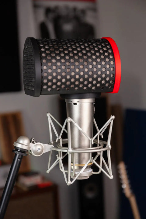 A close-up of the Alpaca Mic condenser microphone with a pop filter, mounted on a shock mount and stand in a recording studio, features blurred instruments and acoustic panels for a professional sound background.