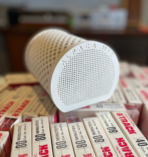 A white, cylindrical Alpaca Mic condenser microphone with a perforated surface sits atop vintage 80 MINUTES audio cassette tapes, suggesting professional sound quality.