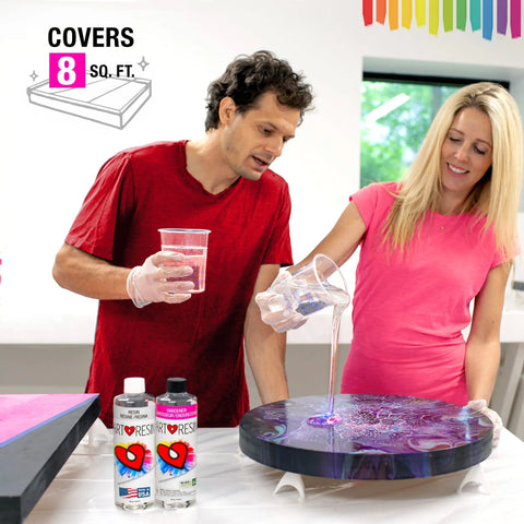 A man and woman wearing gloves pour 32 Oz (946 Ml) ArtResin epoxy resin by ArtResin onto a round artwork, with two resin bottles on the table. A graphic shows Covers 8 sq. ft. beside a tray, and a rainbow stripe decorates the background wall.