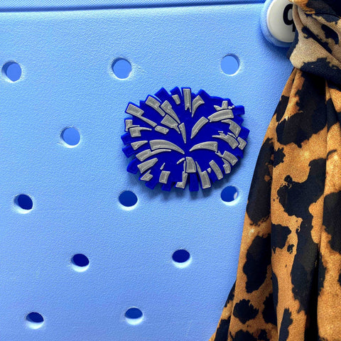 The Chatelet Manufacturing BOGLETS Cheerleading Charm (Blue Pom Pom) is attached to a light blue, hole-punched surface—ideal for customizing Bogg Bags—with a leopard-print fabric beside it for added style.