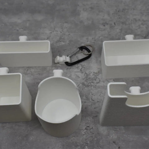 Five white BAGLETS by Chatelet Manufacturing—phone case, key, wallet, carabiner, bottle, and sunglasses holders—are arranged on a gray textured surface. Ideal accessories for Bogg Bags and other totes for organized storage.
