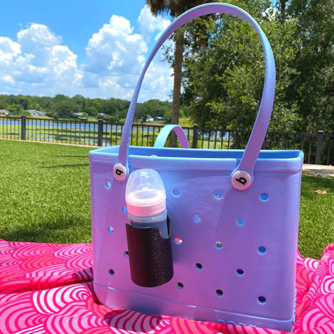A FRESHe BAGLETS Can Drink & Water Bottle Holder Charm Accessory by Chatelet Manufacturing, compatible with Bogg Bags, is attached to a light blue rubber tote bag resting on a pink patterned blanket outdoors under a sunny sky.