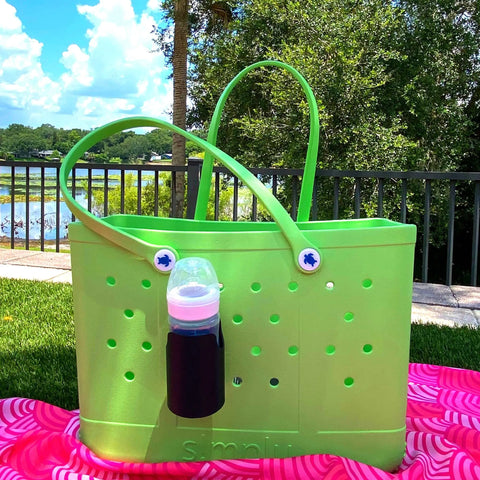 A FRESHe BAGLETS Can Drink & Water Bottle Holder Charm Accessory by Chatelet Manufacturing, compatible with Bogg Bags, hangs on a light green Rubber Tote Bag outdoors on a black fence with greenery, water, blue sky, and a pink patterned towel nearby.