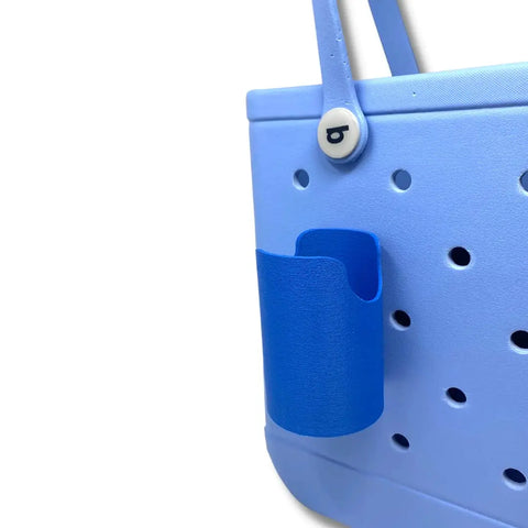 A close-up of a light blue tote bag with a dark blue FRESHe BAGLETS Can Drink & Water Bottle Holder Charm Accessory by Chatelet Manufacturing, compatible with Bogg Bags, attached to its side, plus a round button with the letter b near the handle.