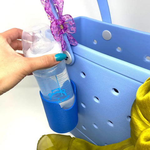 A hand with blue nails places a baby bottle into the Chatelet Manufacturing FRESHe BAGLETS Can Drink & Water Bottle Holder Charm Accessory, attached to a blue Bogg Bag with holes and a purple butterfly clip, with yellow fabric peeking out for a playful touch.