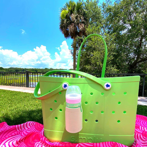 A FRESHe BAGLETS Can Drink & Water Bottle Holder Charm Accessory by Chatelet Manufacturing, compatible with Bogg Bags, is featured outdoors on a pink patterned blanket among grass, trees, and a blue sky with clouds.