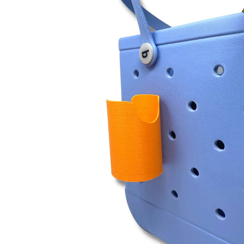 A FRESHe BAGLETS Can Drink & Water Bottle Holder Charm Accessory by Chatelet Manufacturing is attached to a light blue perforated rubber tote, keeping your small orange drink can or water bottle handy and secure on the side.