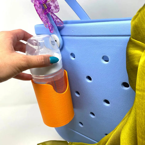 A hand places a baby bottle into an orange FRESHe BAGLETS Can Drink & Water Bottle Holder Charm by Chatelet Manufacturing, attached to a light blue tote bag with holes. A green fabric and purple clip are also visible on the bag.