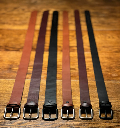 Six Rogue Industries Chamberlain Leather Belts (1.25 wide) in brown and black are displayed side by side on wood, each handcrafted in the USA with a durable metal buckle facing forward.