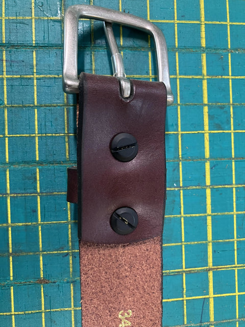 A close-up of the Rogue Industries Chamberlain Leather Belt, 1.25 wide, featuring a silver buckle and two black Chicago screws, shown on a green cutting mat with a yellow grid.