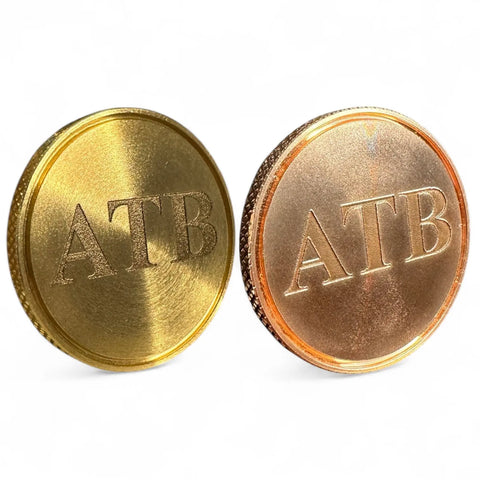 Two upright coins, gold and copper-colored, display “ATB” on a white surface—Fairway Reserve’s Personalized Ball Markers (Set of 2) are stylish brass and copper accessories for your next golf game.
