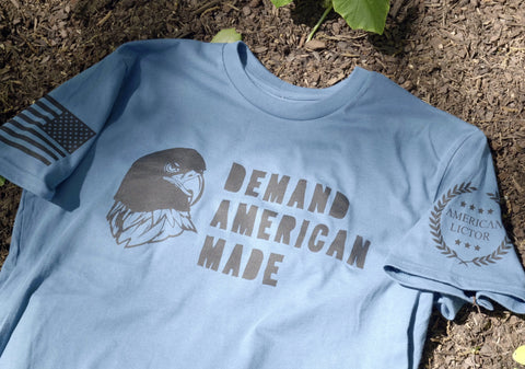 The American Lictor “Demand American Made” T-shirt in light blue showcases a bold bald eagle and slogan on the front, with a U.S. flag on one sleeve and “American Lictor” plus laurel leaves on the other. See our Size Guide for your perfect fit.