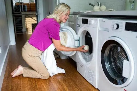 A woman smiles as she uses the Reusable Dryer Ball Set Natural Wool by American Blossom Linens, placing the wool dryer balls into her front-loading washing machine to make laundry day easier—no need for fabric softeners.
