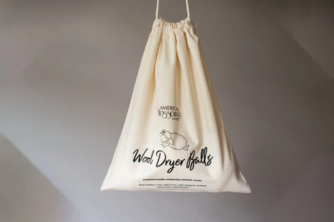 A beige cotton drawstring bag by American Blossom Linens hangs against a gray background. Labeled Wool Dryer Balls with a sheep design, it holds the Reusable Dryer Ball Set Natural Wool—a sustainable laundry alternative.