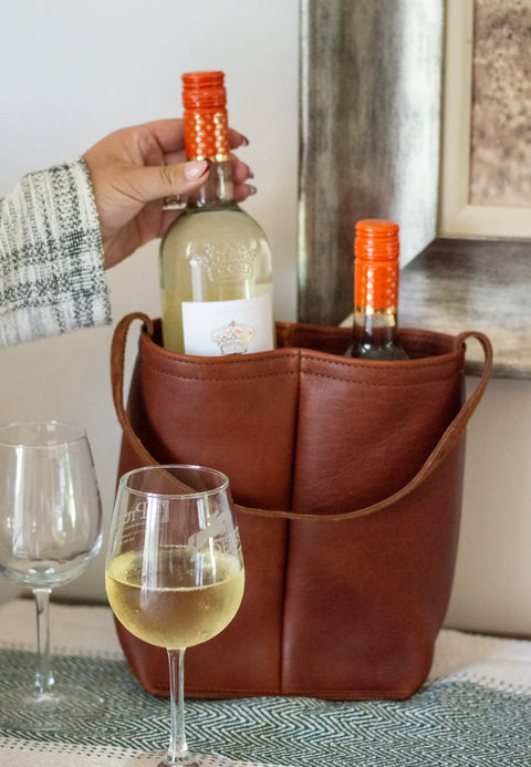 A hand places a white wine bottle with an orange cap into the Urban Southern Urban Dual Wine Tote, a brown full-grain leather carrier already holding another bottle. A full and an empty wine glass rest on a patterned cloth nearby.