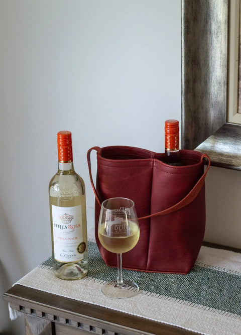 Two bottles of Stella Rosa Stella Peach wine—one in an Urban Southern Urban Dual Wine Tote and one beside it—rest on a wooden table, with a glass of white wine in front near a wall mirror.