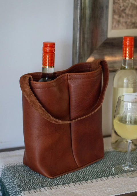 The Urban Southern Urban Dual Wine Tote, crafted from full-grain leather, holds two wine bottles—one with an orange cap. Nearby are a glass of white wine and another bottle on a woven table runner.