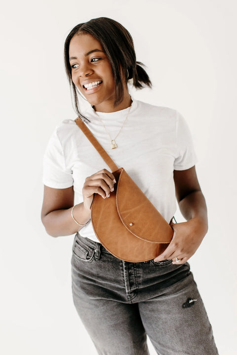 A smiling woman in a white t-shirt and dark jeans holds the Urban Southern Half-Moon Belt Bag, a tan leather fanny pack handcrafted in the USA, across her chest. She stands against a plain white background.